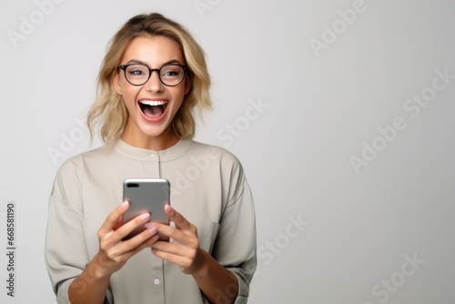 Portrait of excited and surprised young woman looking at mobile phone happy, laughing amazed at message, standing against white background