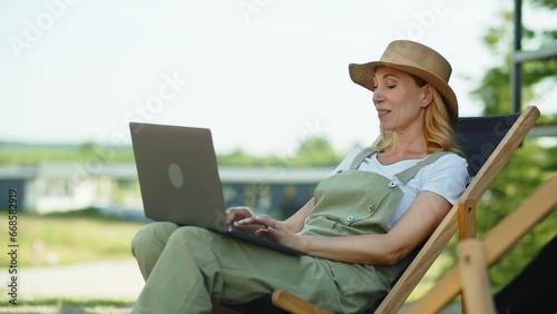 A charming middle aged woman spends her vacation in the countryside. A blogger uses laptop, maintains active correspondence with subscribers, sitting in a wooden recliner. A smiling lady in pleased
