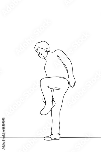 man stands on one leg adjusting his shoes or the bottom of his pants - one line art vector. concept man stopped to check clothes