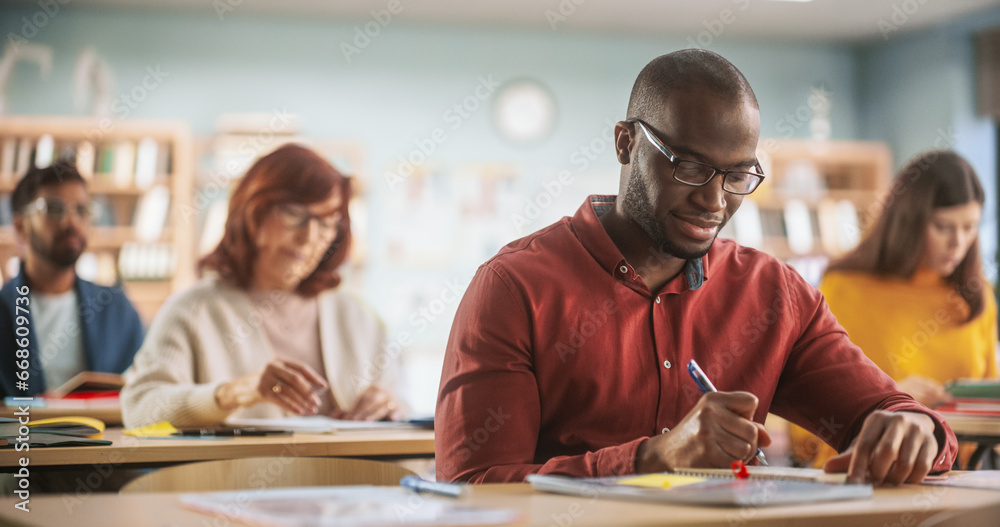 Fototapeta premium Young Black Man Motivated to Learn New Skills During an Adult Education Training Course in School. Diverse Mature Students Learning in Classroom, Reading Textbooks and Writing in Their Notebooks.