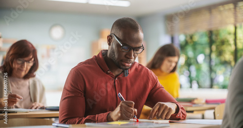 Handsome African Man Writing Down Notes in Notebook. Group of International Mature Students Studying in Classroom. Modern Adult Training Center Help People to Develop New Useful Skills Throughout Life photo