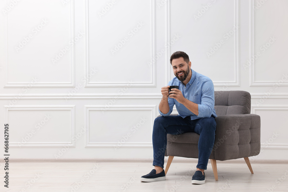 Handsome man with cup of drink sitting in armchair near white wall indoors, space for text