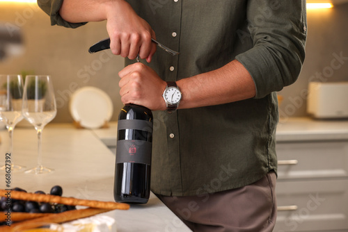 Romantic dinner. Man opening wine bottle with corkscrew at countertop in kitchen, closeup