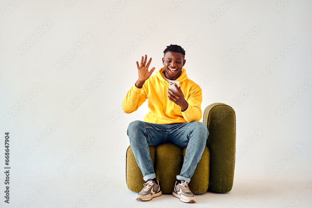 Gadgets for online talks. Happy man in yellow hoodie having video call on smartphone while sitting on green design chair. African guy waving hand to cell phone webcam over white studio background.