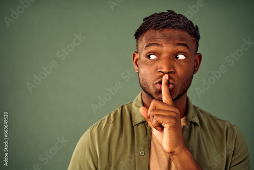 Keep silence, shh. Handsome african american dressed in unbuttoned khaki shirt holding forefinger near lips and looking aside while asking for silence. Isolated over green background. Copy space. photo