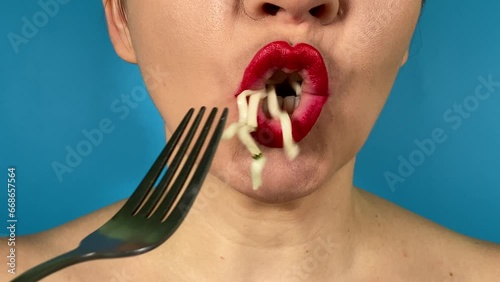Unrecognizable young adult woman vibrantly colored lips eating noodles using a fork, having delicious lunch indoors. Close-up mouth red lipstick savoring pasta or ramen isolated on blue background
