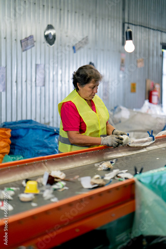 A serious older woman works at waste recycling station and sorts garbage for further processing