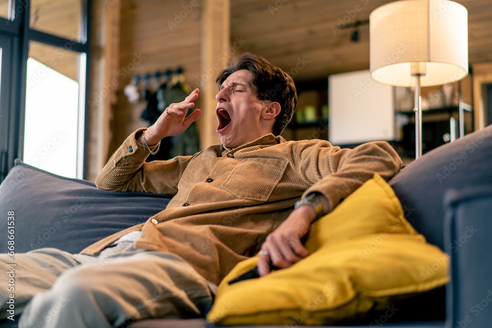 Obraz premium A young guy yawns sitting on the sofa with remote control in his hand watching TV at home