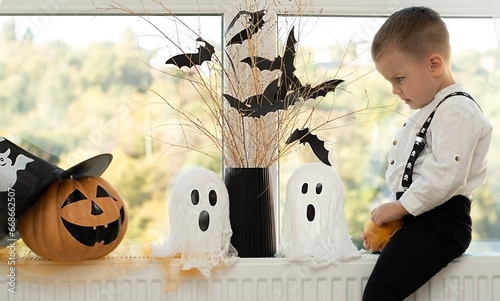 Halloween. Concept. A cute and cheerful boy sits near the window against the background of white ghosts and a vase with dry branches and black paper bats.