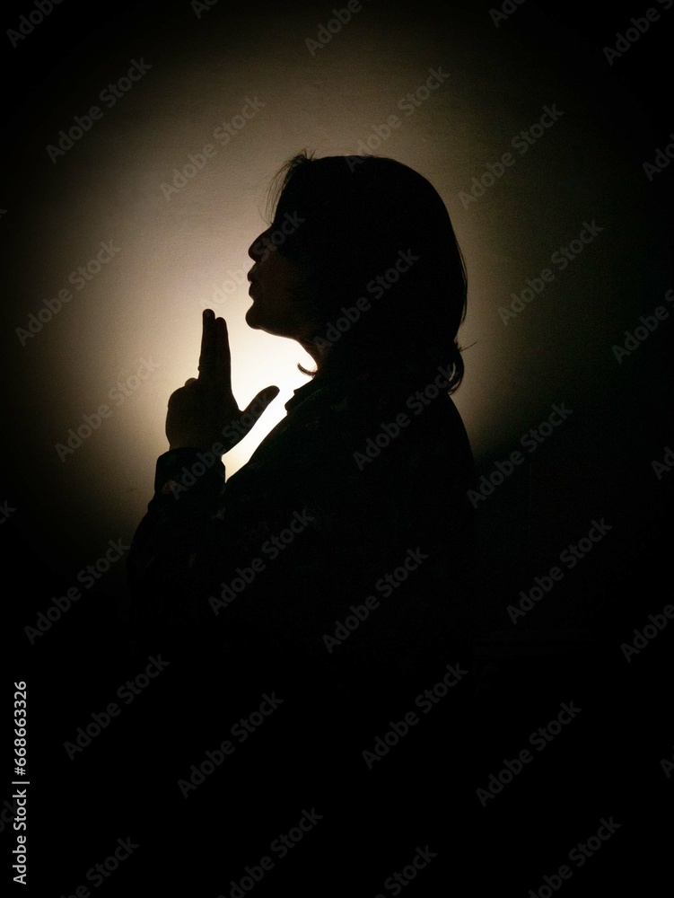 A beautiful silhouette of a young plump woman in the dark, holding her hands with a gun up, lips stretched forward. Shadow, religious rituals. Round lighting, contour light