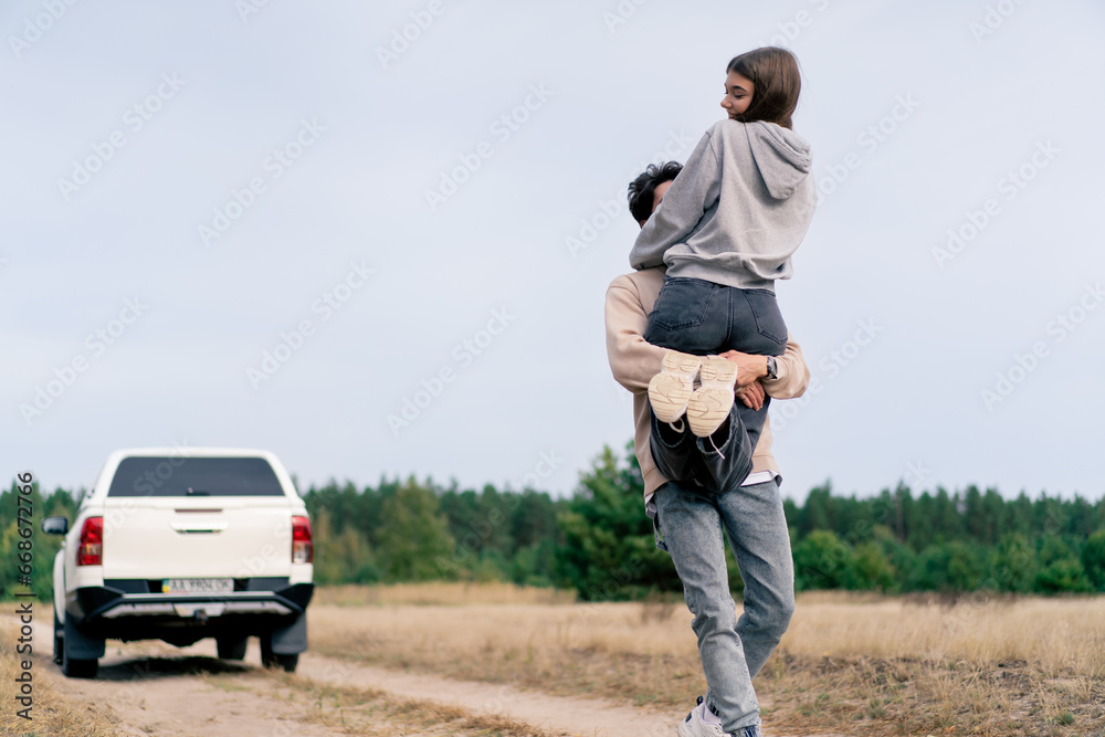 A young guy spins his girlfriend in his arms during stop in forest steppe while traveling by car