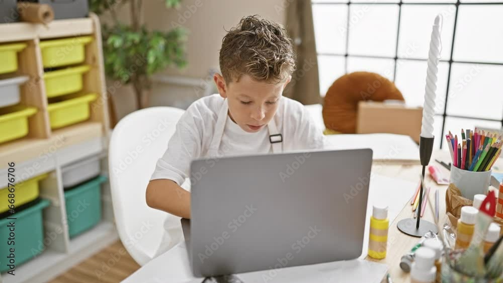 Adorable blond artist boy seriously concentrating on laptop at art studio, sitting in a cute apron, immersed in online painting lesson Adorable blond artist boy seriously concentrating on laptop at art studio, sitting in a cute apron, immersed in online painting lesson