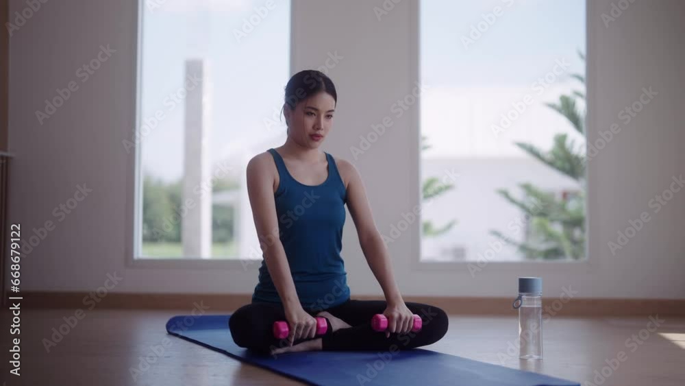 An Asian woman in workout attire is using dumbbells to strengthen her muscles for good health and strength on a yoga mat for home workouts An Asian woman in workout attire is using dumbbells to strengthen her muscles for good health and strength on a yoga mat for home workouts