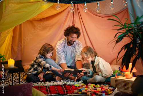 A good looking dad with curly hair is spending his time with his two young boys bonding and reading a fun book together as they are all in a very big handmade blanket fort indoors