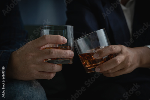 Close-up view of two men in formal attire clinking whiskey glasses, stylish businessman friends in suits toasting with whiskey glasses in home, closeup