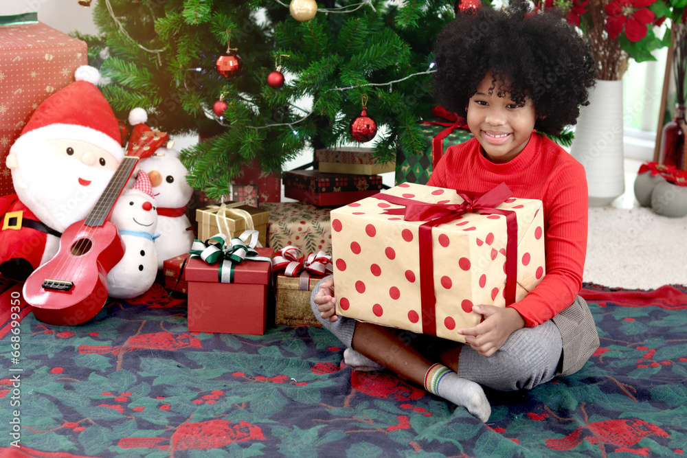 Portrait of happy smiling African kid girl with curly hair holding gift box present while sitting under decorative Christmas tree. Child celebrates winter holiday. merry Christmas, happy childhood Portrait of happy smiling African kid girl with curly hair holding gift box present while sitting under decorative Christmas tree. Child celebrates winter holiday. merry Christmas, happy childhood