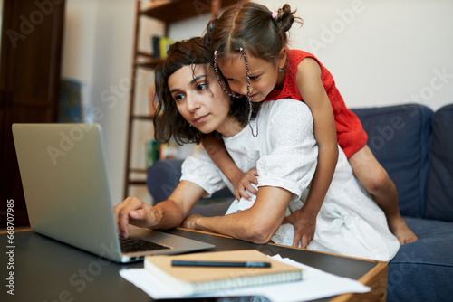 Working mother. Young mom freelancer sitting in front of laptop while her daughter climbing on her back to play together, disturbing her from important work, female combining business and motherhood