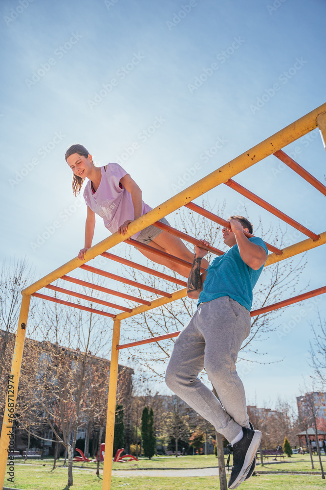 Handsome male sports person doing pull ups in the park while his female friend is doing push ups on horizontal metal bars