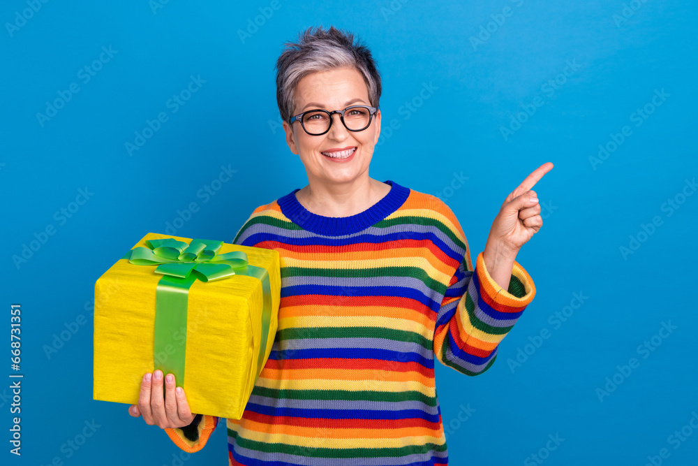 Photo of cheerful nice aged lady toothy smile hold giftbox indicate finger empty space isolated on blue color background Photo of cheerful nice aged lady toothy smile hold giftbox indicate finger empty space isolated on blue color background