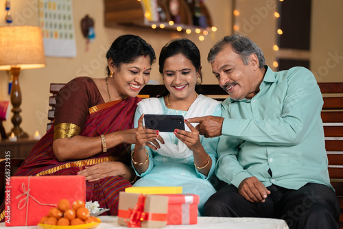 Joyful happy Indian mother and father watching mobile phone with daughter at home during diwali festival celebration - concept of cyberspace  social media sharing and online app streaming