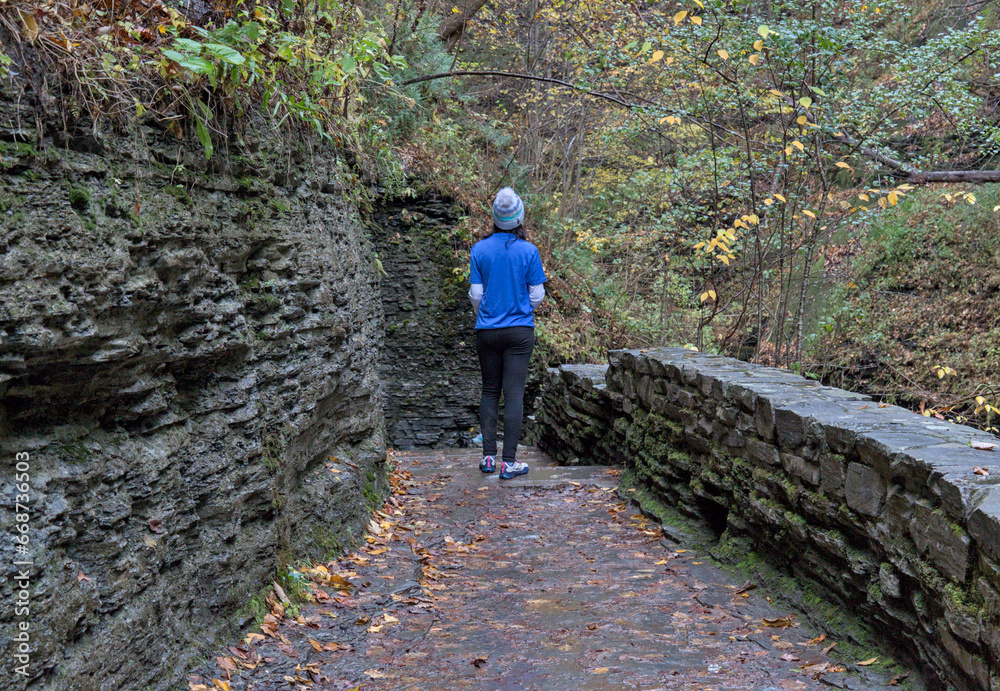 woman walking on a trail in watkins glen state park (shot from behind, unrecognizable) wearing bright blue t-shirt, winter hat, dark pants (autumn, leaves changing colors, gorge waterfall trail hike)