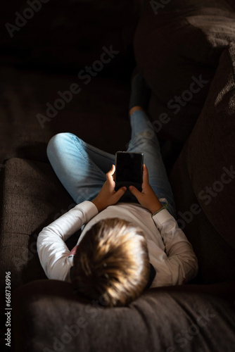 A child lying on the couch, staring at a mobile phone. Addiction and dependence of children and youth on new technologies. Parents' concern about children's obsession with consuming online content