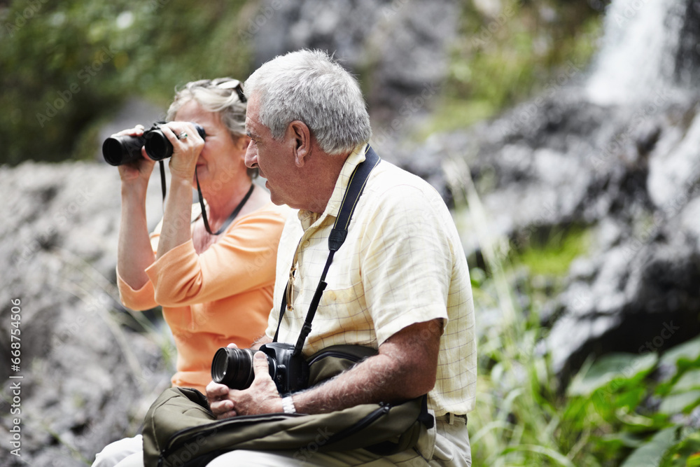 Senior, couple and binocular in forest for tourist, sightseeing or vacation with waterfall and scenery. Elderly, man and woman in woods or nature for holiday, adventure or experience with backpack