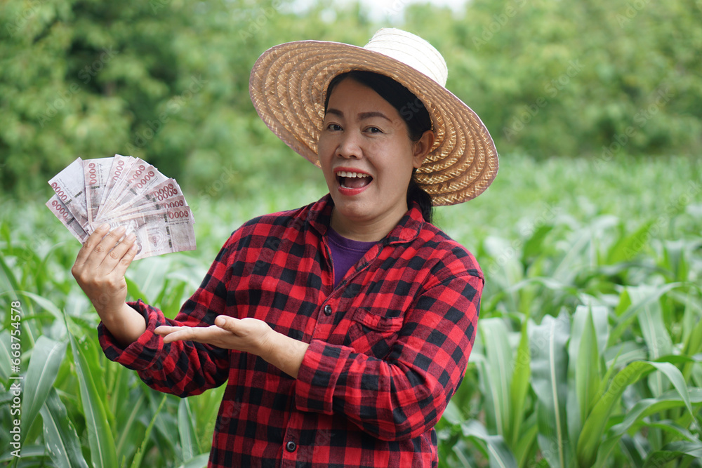 Asian woman farmer is at garden, wears hat. red plaid shirt, holds Thai notebank money. feels excited. Concept, agriculture occupation. Thai farmer lifestyle. Satisfied to get income or profit. 