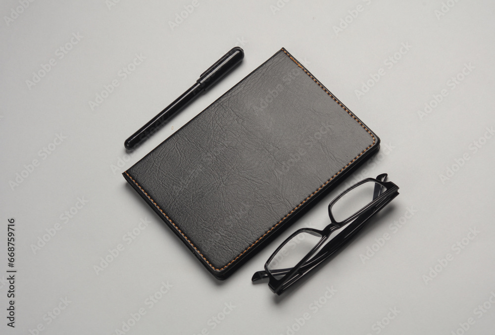 Notebook with black leather cover and pen, eyeglasses on gray background. Business concept. Notebook with black leather cover and pen, eyeglasses on gray background. Business concept.