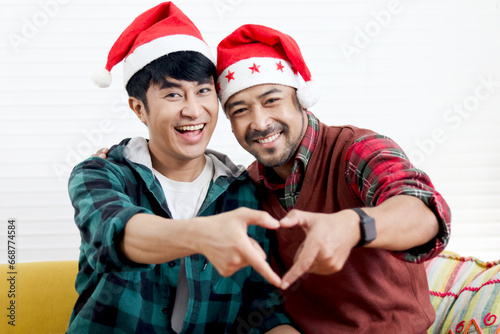 Cute happy young LGBT couple wearing Sata hat, making heart shape by hands on sofa in living room. Asian gay male lover sharing romantic special moment on Christmas holiday together.