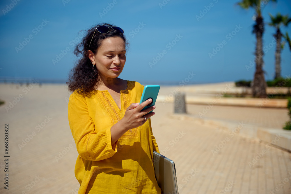 Happy woman checking mobile app on smartphone, holding laptop in hand while strolling the promenade, going to the office