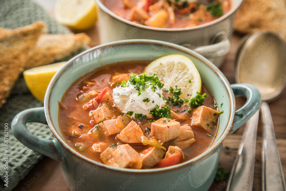 Ost Deutsche DDR GDR Soljanka Eintopf Suppe mit Jagdwurst, Saure Gurken, Letscho, Schmand, Zitrone, Toast und Petersilie auf Holzbrett mit Löffel, Salzstreuer und hell grau Stein Hintergrund Ost Deutsche DDR GDR Soljanka Eintopf Suppe mit Jagdwurst, Saure Gurken, Letscho, Schmand, Zitrone, Toast und Petersilie auf Holzbrett mit Löffel, Salzstreuer und hell grau Stein Hintergrund