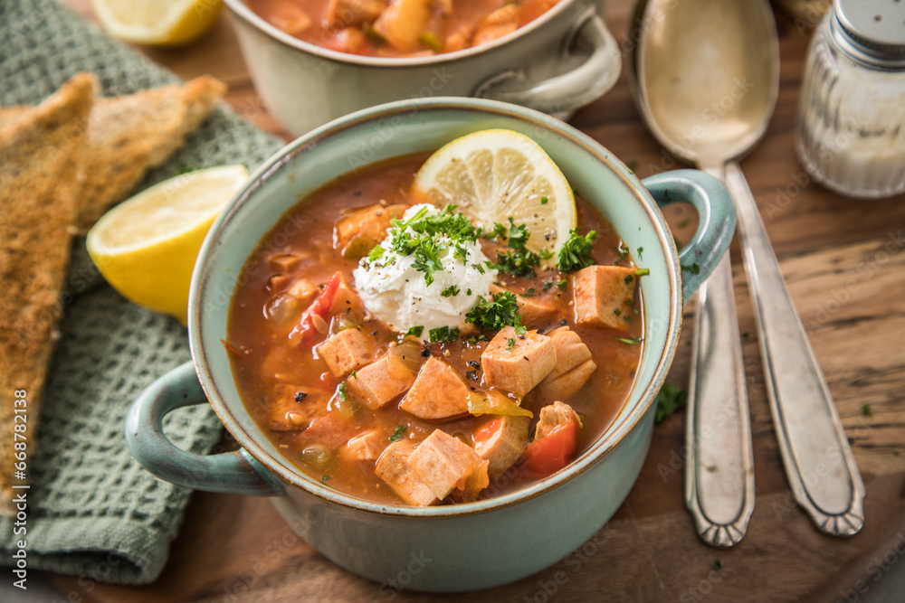 Ost Deutsche DDR GDR Soljanka Eintopf Suppe mit Jagdwurst, Saure Gurken, Letscho, Schmand, Zitrone, Toast und Petersilie auf Holzbrett mit Löffel, Salzstreuer und hell grau Stein Hintergrund Ost Deutsche DDR GDR Soljanka Eintopf Suppe mit Jagdwurst, Saure Gurken, Letscho, Schmand, Zitrone, Toast und Petersilie auf Holzbrett mit Löffel, Salzstreuer und hell grau Stein Hintergrund