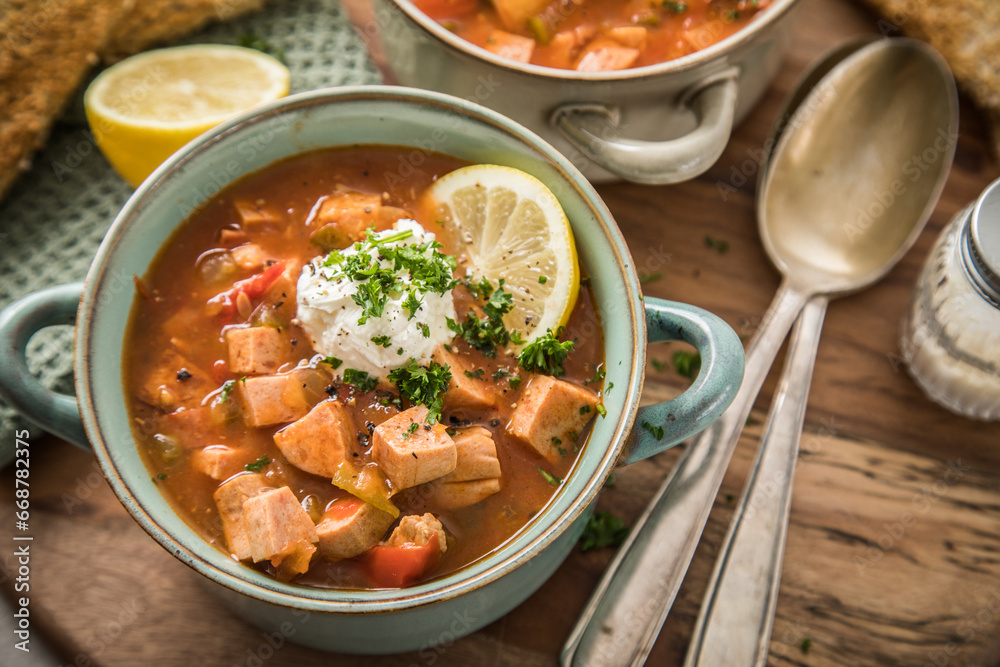 Ost Deutsche DDR GDR Soljanka Eintopf Suppe mit Jagdwurst, Saure Gurken, Letscho, Schmand, Zitrone, Toast und Petersilie auf Holzbrett mit Löffel, Salzstreuer und hell grau Stein Hintergrund