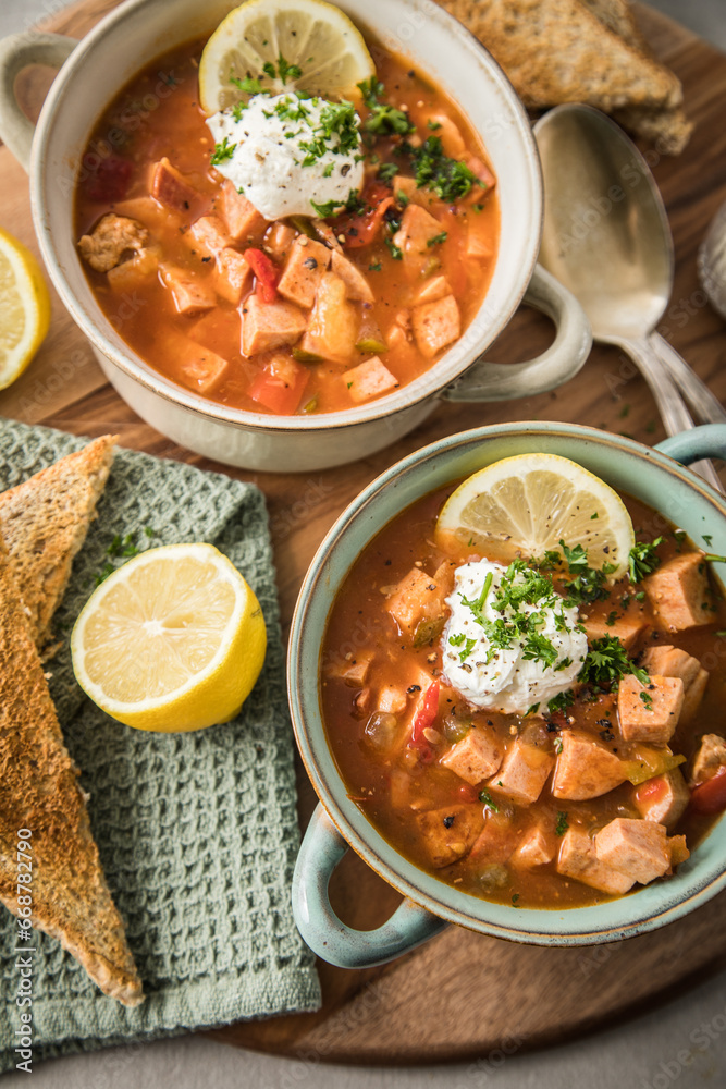 Ost Deutsche DDR GDR Soljanka Eintopf Suppe mit Jagdwurst, Saure Gurken, Letscho, Schmand, Zitrone, Toast und Petersilie auf Holzbrett mit Löffel, Salzstreuer und hell grau Stein Hintergrund Ost Deutsche DDR GDR Soljanka Eintopf Suppe mit Jagdwurst, Saure Gurken, Letscho, Schmand, Zitrone, Toast und Petersilie auf Holzbrett mit Löffel, Salzstreuer und hell grau Stein Hintergrund