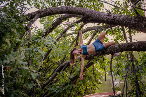 An attractive fit girl exercises outdoors in a park, climbing a tree and training with an elastic. She stretches and warms up, showcasing her flexibility and strength.