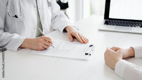 Doctor and patient sitting near each other at the white desk in clinic. Female physician is listening filling up a records form. Medicine concept