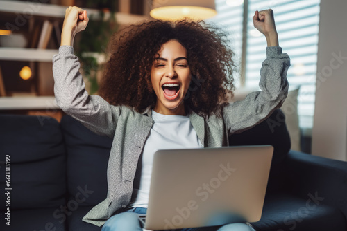 Beautiful young multiethnic woman smiling and rejoices after success. Happy woman celebrating business success on sofa in living room with computer.