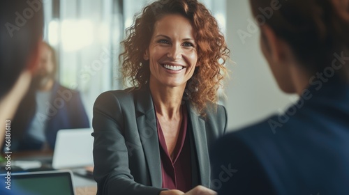 An upbeat, middle-aged businesswoman who is a manager is seen shaking hands during an office meeting. She's greeting a smiling female HR professional conducting a job interview photo