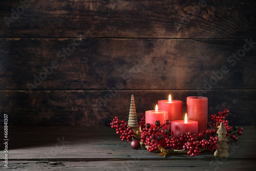 Third advent wreath with red candles, three are lit, decoration with berries, Christmas balls and small wooden trees, dark rustic background, copy space, selected focus