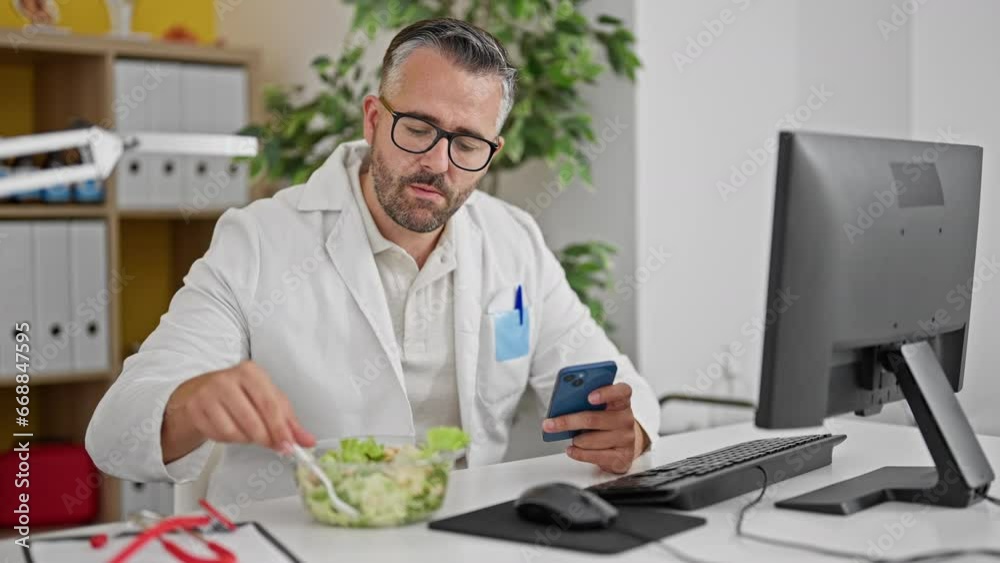 Grey-haired man doctor eating salad using smartphone at the clinic