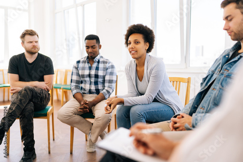 Low-angle view of African alcohol addict female sharing anger management issues with people at aa rehab meeting. Stressed young woman with mental health problems talking to patients and psychologist.