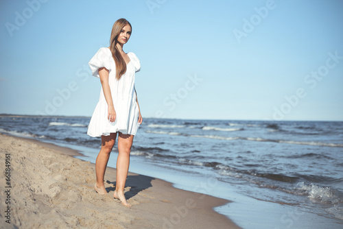 Happy smiling beautiful woman is walking on the ocean beach in a white summer dress © rogerphoto