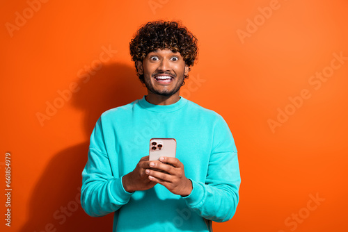 Photo of young surprised funny positive man indian tik tok user wearing teal pullover using smartphone isolated on orange color background