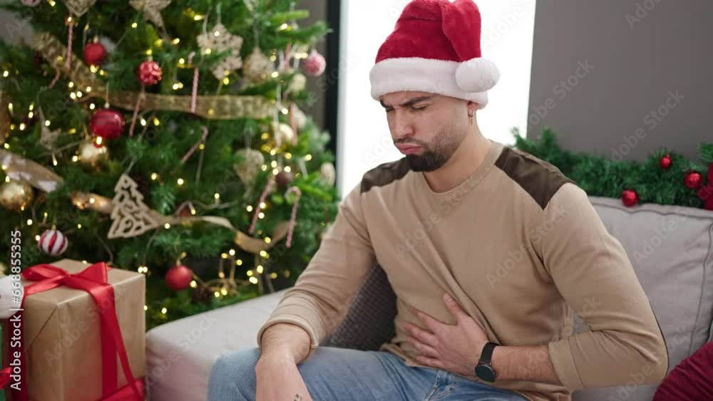 Young arab man suffering stomach ache sitting by christmas tree at home Young arab man suffering stomach ache sitting by christmas tree at home