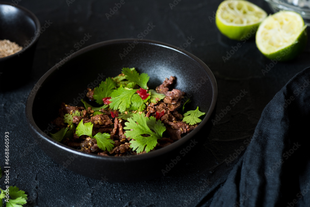 Spicy carpaccio salad with green coriander, lime, sesame seeds and red hot chili pepper in a black bowl.