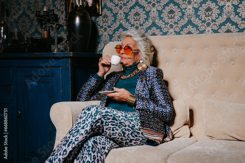 Elegant senior woman in trendy and luxury wear drinking coffee while sitting on the couch at home