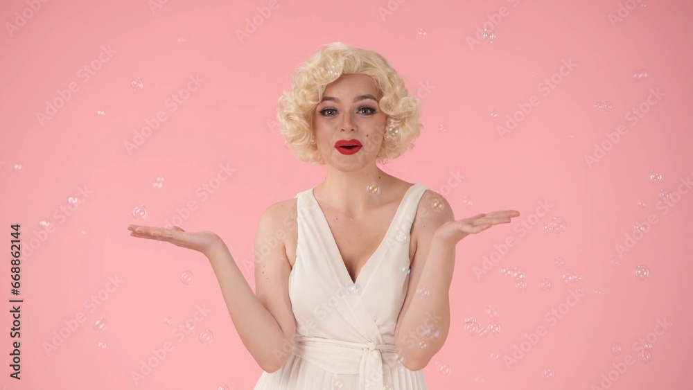 Cheerful beautiful young woman surrounded by soap bubbles. Woman looking like Marilyn Monroe in studio on pink background. Emotions, joy, childhood. Cheerful beautiful young woman surrounded by soap bubbles. Woman looking like Marilyn Monroe in studio on pink background. Emotions, joy, childhood.
