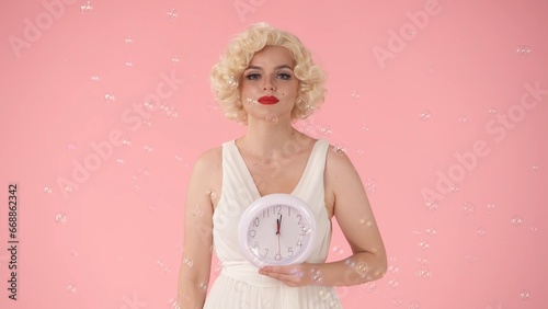Woman holding a white round wall clock in her hand. Woman in image on Marilyn Monroe, in studio surrounded by soap bubbles on pink background. Concept of time.
