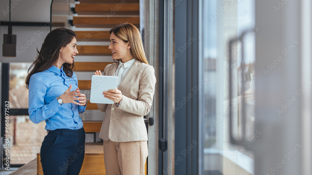 Fototapeta premium Two business women having a discussion, they're standing in an office and using a tablet. Professional women making a to do list as part of their project 